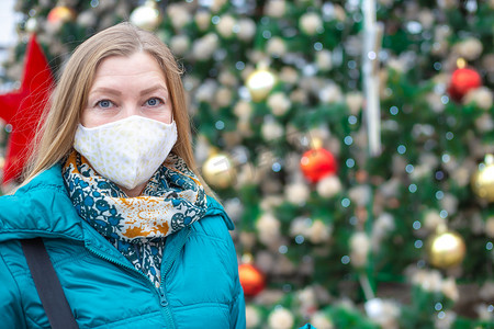 街上戴口罩的人摄影照片_一名戴着防护面具的妇女站在街上一棵装饰得节日的圣诞树旁。