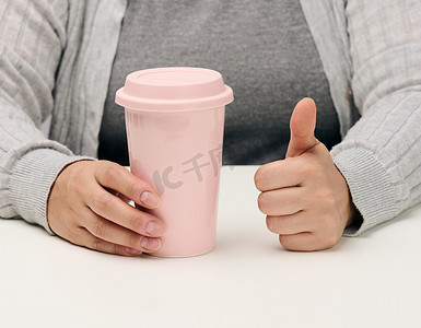 穿粉色衣服的女人摄影照片_穿灰色衣服的女人拿着一个粉红色的陶瓷杯，女人坐在一张白色的桌子旁，一天的开始，早上