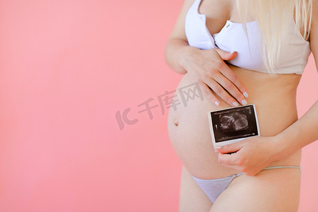 年轻怀孕女孩抱着腹部在粉红色单声道背景下与超声波。