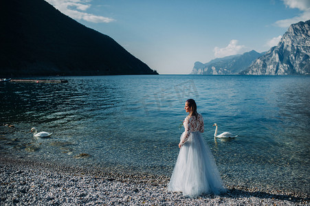 加衣服摄影照片_一个穿着漂亮白色连衣裙的女孩沿着加尔达湖的堤岸行走。一名妇女在意大利的山和湖的背景下被拍照。托尔博莱