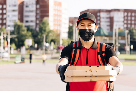 年轻的男性食品快递员戴着防护面具，背着绝缘的红色背包走在城市街道上运送订单。
