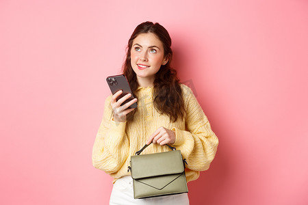 美丽的女模特拿着钱包和智能手机，看起来很体贴，思考消息的最佳答案，站在粉红色的背景下