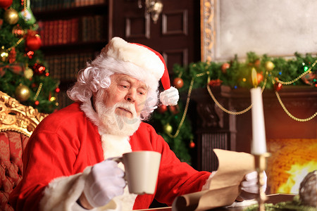 快乐的圣诞老人坐在圣诞树附近的房间里阅读圣诞信件或愿望清单的肖像。