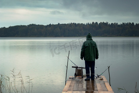 钓鱼竿摄影照片_雨天独自一人的渔夫站在木桥上用鱼竿钓鱼