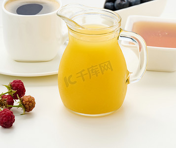 鲜榨果汁摄影照片_鲜榨果汁装在玻璃透明醒酒器中，一杯咖啡，白色桌子上的蜂蜜。