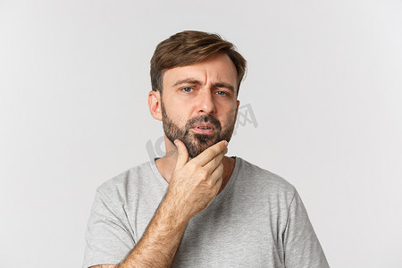 困惑脸摄影照片_困惑的男人摸着胡子，看着自己，需要刮胡子，站在白色背景上的特写