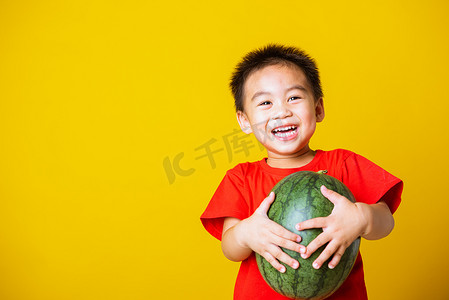 快乐的亚洲儿童或孩子可爱的小男孩穿着红色T恤，笑容迷人，玩耍时手里拿着未切的完整西瓜