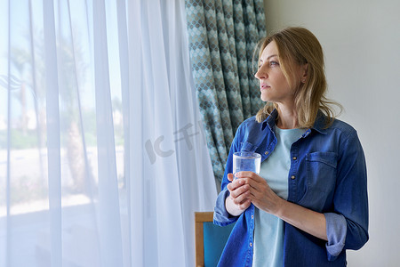 中年妇女拿着一杯水站在家里靠近窗户的地方，复印空间