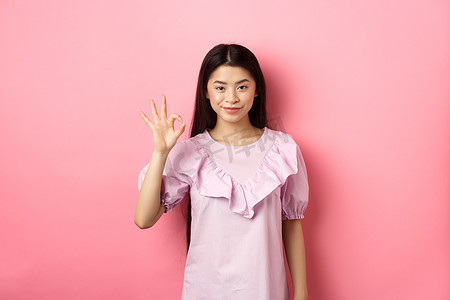穿着裙子的微笑亚洲女性表现出良好的迹象，赞美和赞美好东西，批准优秀的选择，站在粉红色的背景上