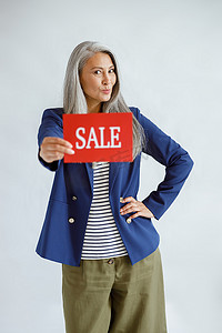 夹克里头发灰白的成熟亚洲女士在工作室里拿着写着“销售”字样的红卡