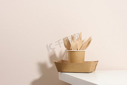 桌子上的空盘子摄影照片_纸纸板棕色盘子和杯子，白色桌子上的木叉和刀，米色背景。