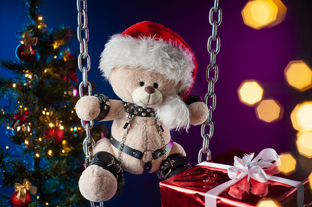 戴着圣诞老人帽子的泰迪熊是圣诞树背景上 BDSM 游戏的圣诞礼物