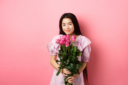浪漫温柔的亚洲女孩拿着玫瑰花束，对着镜头微笑可爱，与情人约会，穿着裙子，粉红色背景