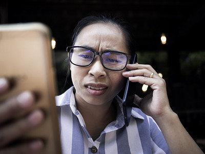 咖啡厅女孩摄影照片_在咖啡厅打电话时，压力重重的亚洲年轻女性拿着智能手机上班，脸上不快乐而严肃。