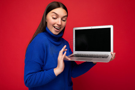 照片中，美丽的黑发年轻女子拿着电脑笔记本电脑，穿着蓝色毛衣，在红墙背景下俯视