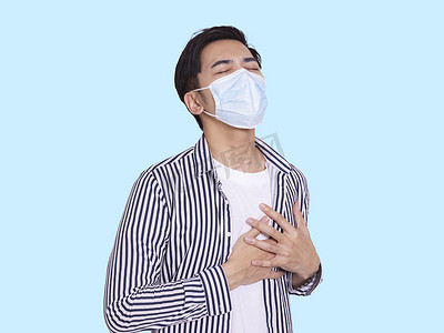 性疾病摄影照片_戴着防护性医用口罩的年轻人，双手放在胸前感到不舒服，呼吸困难。