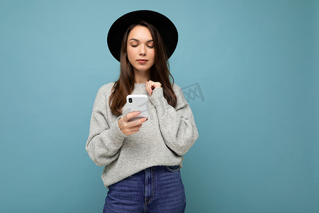 美丽的黑发年轻女子戴着黑帽子，穿着灰色毛衣，拿着智能手机，低头看着背景中孤立的电话短信