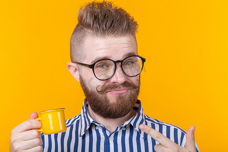 可爱的小杯子摄影照片_一个有趣、自信、英俊的年轻人的肖像，戴着眼镜，留着胡子，黄色背景上有一个黄色小杯子。
