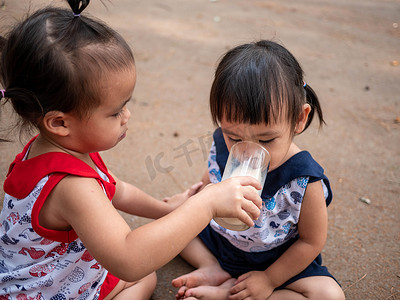 亚洲小孩坐在花园里，用玻璃杯给姐姐喂牛奶