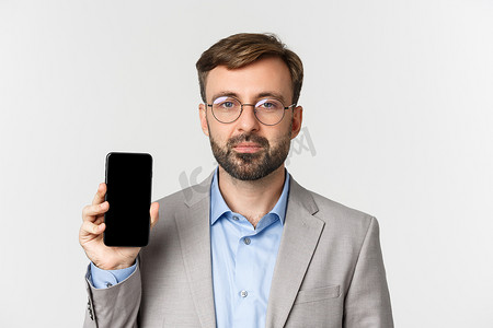 公司网页背景摄影照片_戴着眼镜和灰色西装的英俊商人的特写镜头，显示手机屏幕、演示应用程序或网页，站在白色背景上
