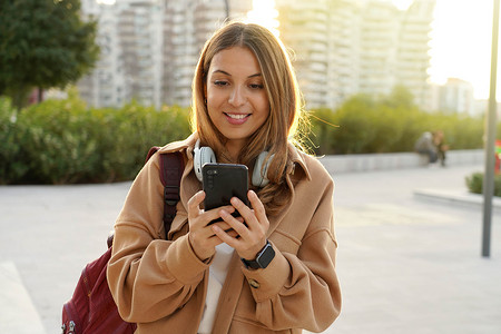 日落时分，穿着外套和背包的阳光女孩在现代城市街道上与智能手机应用程序聊天。