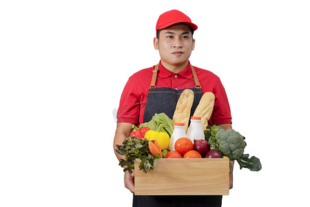红色长围裙摄影照片_身穿红色制服的杂货送货快递员，手里拿着装有食物、新鲜水果和蔬菜的杂货盒。