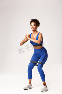蓝色运动服摄影照片_全长微笑的适合女性享受健身，穿着蓝色运动服，用阻力带蹲下，站在白色背景下