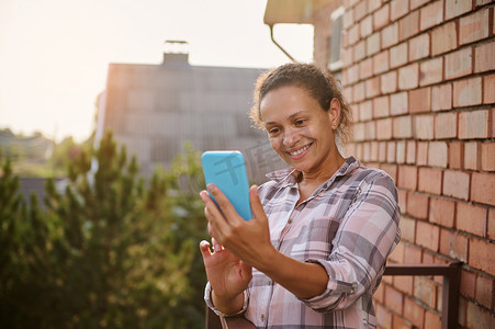 阳光明媚、温暖的夏日，快乐的女人用智能手机站在乡村别墅的阳台上自拍