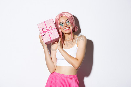 盒子粉色摄影照片_戴着粉色假发的漂亮女孩拿着生日礼物摇晃着盒子，想知道包裹着的盒子里装着什么，站在白色背景上