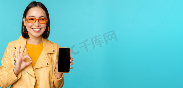 微笑的韩国女性展示手机应用程序界面、智能手机应用程序、手机推荐、站在蓝色背景上