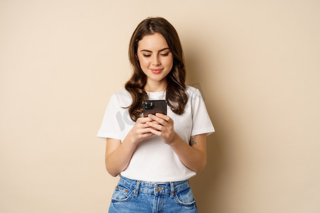 年轻的现代女孩在应用程序上聊天，使用智能手机应用程序，微笑着，站在米色背景上