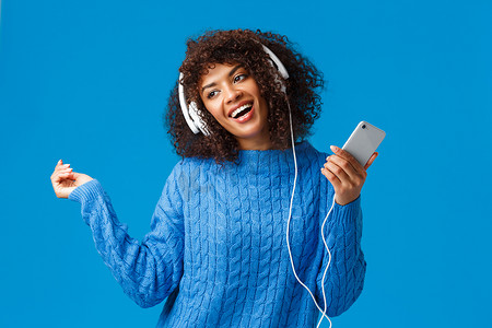 快乐无忧无虑的可爱非洲裔美国女模特穿着冬季毛衣，随着节奏音乐跳舞，在歌曲应用程序上听新曲目，圣诞播放列表，戴耳机拿着智能手机