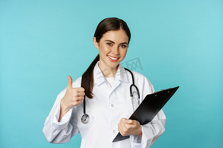 自信的微笑医生女医生，医生竖起大拇指，拿着剪贴板，在医院预约，站在绿松石背景上