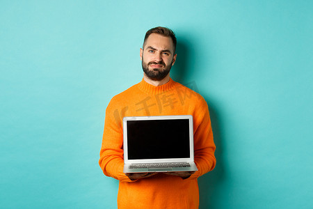 穿着橙色毛衣、留着胡子的帅哥展示笔记本电脑屏幕，展示促销品，站在浅蓝色背景上，表情失望、心烦意乱
