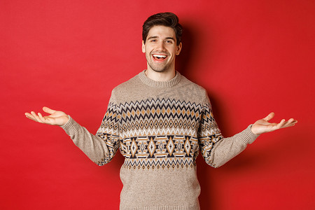 一双手拿东西摄影照片_庆祝新年假期的快乐帅哥的肖像，穿着圣诞毛衣，双手侧向微笑，在复印空间上拿着一些东西，站在红色背景上