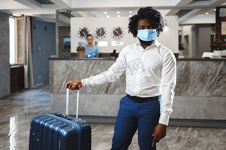 办理摄影照片_带着手提箱的非洲男子酒店客人戴着防护面罩，以防止感染冠状病毒