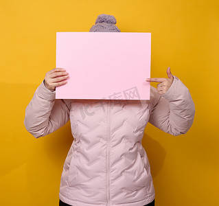 穿着粉色冬季夹克和帽子的女人拿着黄色背景上的空白粉色纸。