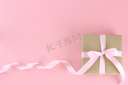 顶视图棕色纸礼盒，在柔和的粉红色背景上配有粉红色缎面卷曲丝带蝴蝶结。