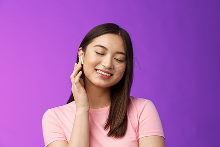 紫色背景音乐摄影照片_可爱的亚洲女孩享受听音乐，戴上无线耳机，倾斜头闭上眼睛梦幻般的微笑满意，完美的声音，使音量耳机更大，站立紫色背景
