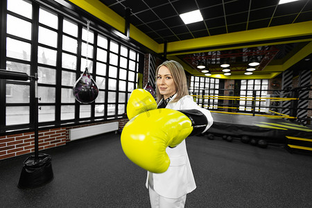 戴着黄色拳击手套的坚定、时尚的女商人在拳击场的背景下向镜头挥拳