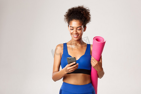 穿着蓝色运动服的年轻运动女性，拿着橡胶垫做瑜伽，使用手机，站在白色背景下