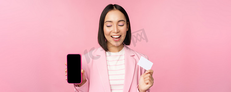 身穿西装的微笑韩国女商人，展示手机屏幕、信用卡、展示网上银行应用界面、粉色背景