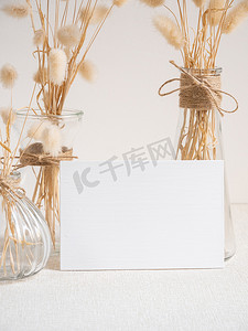 空白白色贺卡模型。米色桌子和水泥墙背景上的现代玻璃花瓶中用干 Lagurus ovatus 花组成的装饰