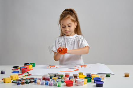穿着白色 T 恤的小女孩坐在桌子旁，上面涂着什么和彩色颜料，正在给她的手涂色。