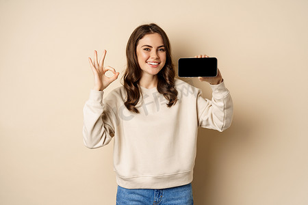 app展示界面摄影照片_微笑的快乐女孩展示智能手机屏幕应用程序、移动界面、好的标志，站在米色背景上