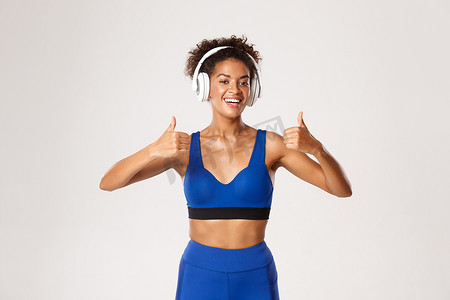 蓝色运动服摄影照片_穿着蓝色运动服的健康运动非洲裔美国女性竖起大拇指，在锻炼时戴耳机听音乐，在白色背景下锻炼
