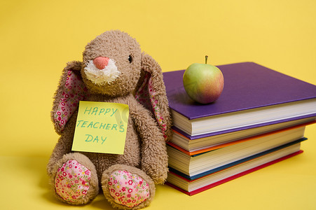 一只毛绒玩具兔子，在黄色便条纸上写着“教师节快乐”，靠在一堆彩色书籍上，黄色背景上有文字空间