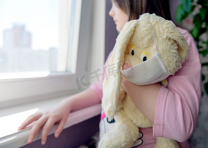 一个带着玩具的女孩，一只毛绒兔子，戴着面具，看着窗外的概念，预防冠状病毒、隔离、待在家里。