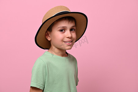 戴草帽的男摄影照片_一个戴着夏季草帽的英俊男学生的侧面肖像，看着相机，站在粉红色的背景上，有复制空间。
