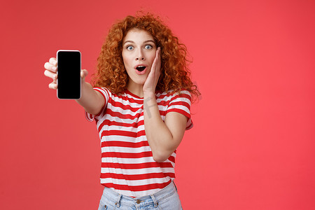 手机app页面摄影照片_有趣的印象深刻的红发好看的卷发兴奋的女孩分享很棒的社交媒体页面显示智能手机屏幕惊讶的触摸脸颊兴奋影响了很棒的应用程序红色背景